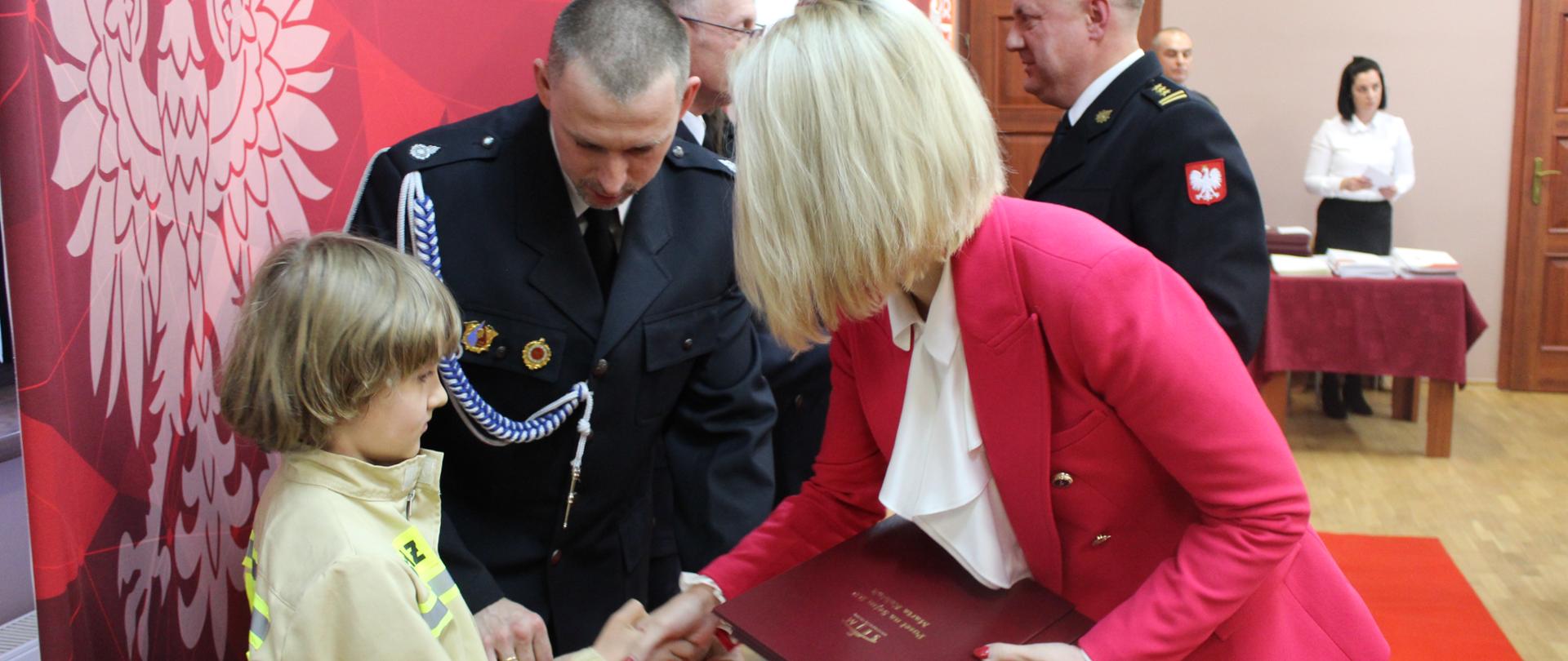 Kobieta blondynka w czerwonej marynarce ściska dłoń dziewczynce ubranej w mundur strażaka, obok strażak mężczyna, w tle grupa ludzi