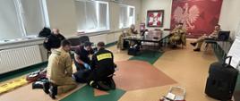Na zdjęciu przedstawiona jest scena z ćwiczeń ratownictwa medycznego w jednostce Państwowej Straży Pożarnej. Kilku strażaków w mundurach zajmuje się osobą leżącą na podłodze, symulującą poszkodowanego. W tle widać inne osoby w mundurach przy stołach, obserwujące ćwiczenia. W sali szkoleniowej znajdują się różne elementy wyposażenia, takie jak nosze i torby medyczne. Na ścianie widnieje czerwony baner PSP z wizerunkiem orła.