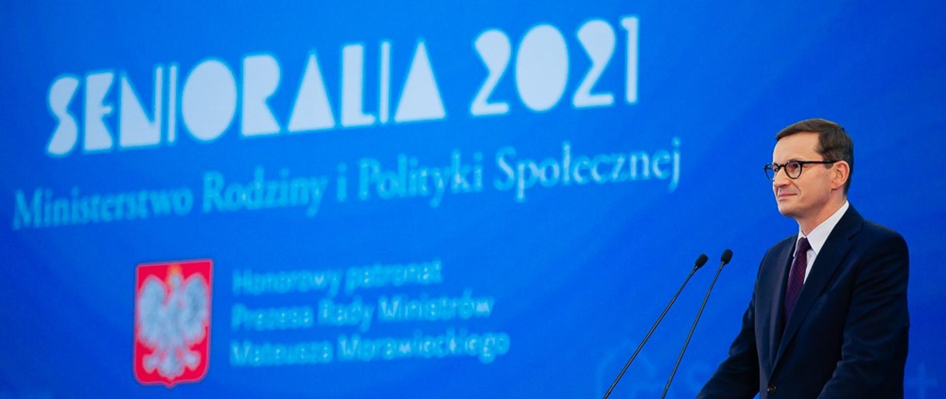 Premier Mateusz Morawiecki przemawia przy mównicy, w tle ścianka multimedialna.