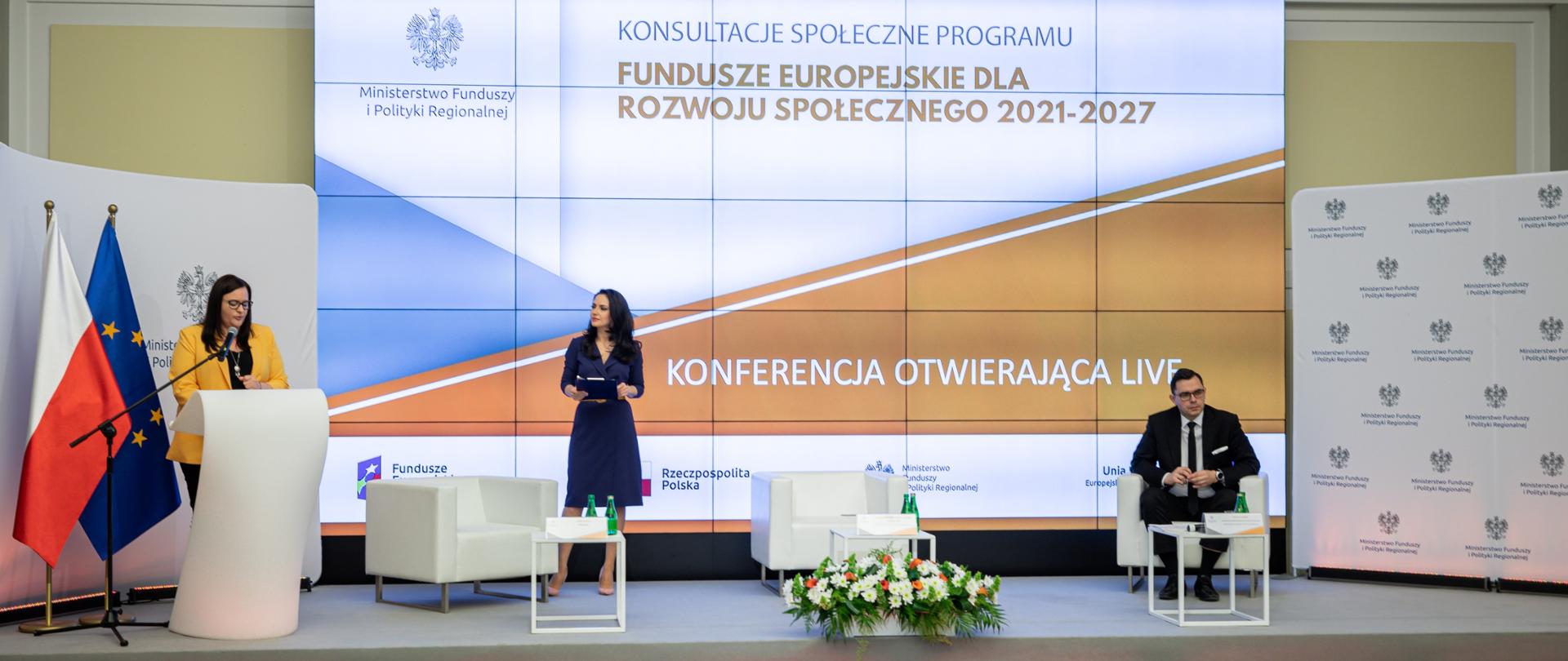 W sali konferencyjnej na scenie są trzy osoby. W mównicy po lewej stronie stoi wiceminister Małgorzata Jarosińska-Jedynak. Obok stoi kobieta. Dalej w fotelu siedzi mężczyzna. Za nimi na ekranie napis: Konsultacje Programu Fundusze Europejskie dla Rozwoju Społecznego 2021-2027, Konferencja otwierająca". Obok mównicy dwie flagi PL i UE.