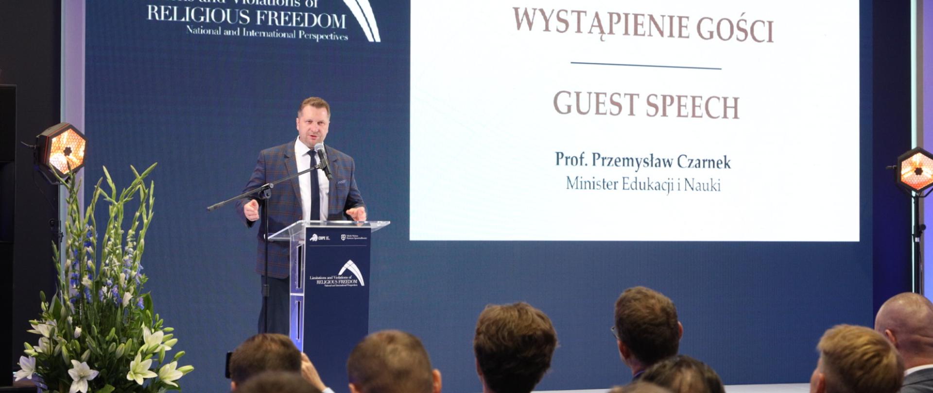 Minister Przemysław Czarnek przemawia stojąc za pulpitem. Za nim w tle plansza z napisem Wystąpienie gości - prof. Przemysław Czarnek Minister Edukacji i Nauki