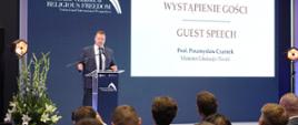 Minister Przemysław Czarnek przemawia stojąc za pulpitem. Za nim w tle plansza z napisem Wystąpienie gości - prof. Przemysław Czarnek Minister Edukacji i Nauki