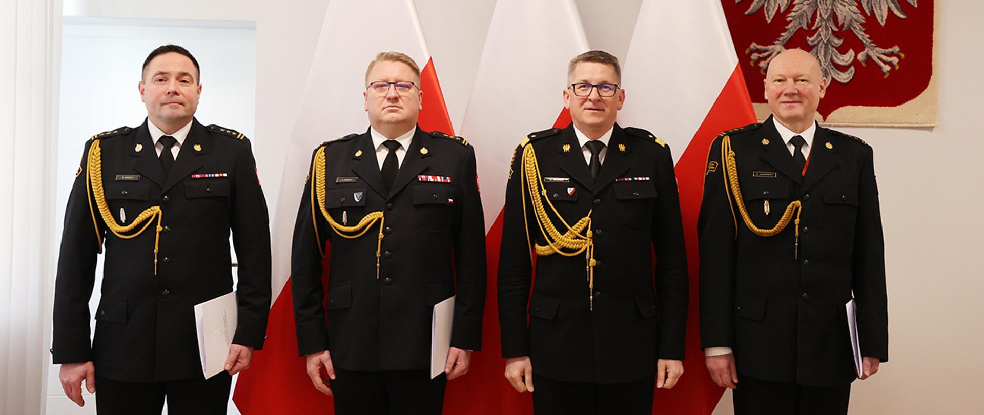 Na tle flag i godła stoją kolejno od lewej: nowy zastępca łódzkiego komendanta wojewódzkiego, łódzki komendant wojewódzki, zastępca komendanta głównego oraz ustępujący zastępca wojewódzki w Łodzi