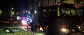 Na zdjęciu w porze nocnej znajdują się dwa wozy strażackie, które stoją przed blokiem. Od samochodów poprowadzone są linie gaśnicze.