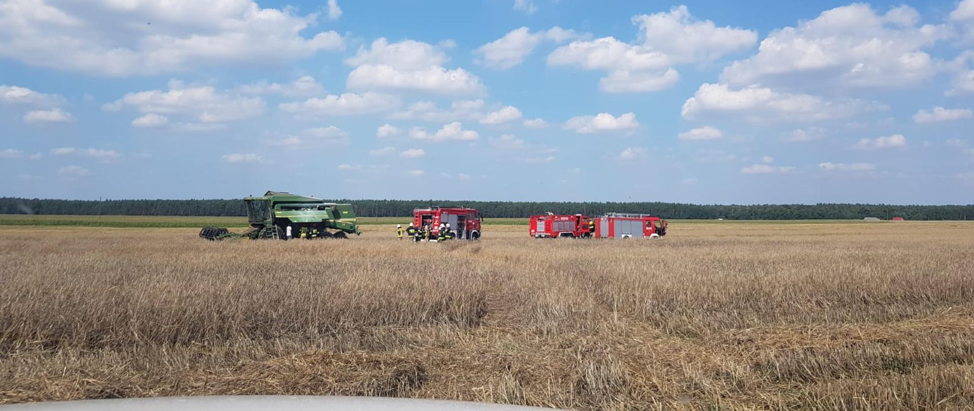 Widoczne miejsce zdarzenia, 3 pojazdy gaśnicze pożarnicze oraz kombajn zbożowy znajdujące się na polu podczas zbiorów. Strażacy sprawdzają miejsce pożaru, ugaszonego przez operatora maszyny przed przybyciem służb. 