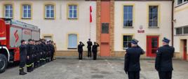 Strażacy w czarnych mundurach stojący w szeregu na utwardzonym placu. Jeden z nich wciąga biało-czerwoną flagę na maszt. W tle czerwono-srebrny strażacki samochód i budynek.