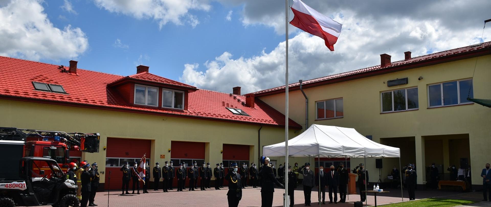 Na zdjęciu widzimy skarżyskich strażaków na placu wewnętrznym Komendy Powiatowej. Poczet flagowy Komendy Powiatowej PSP w Skarżysku-Kamiennej wciąga flagę Państwową na masz. W tle skarżyscy strażacy oraz nieliczni zaproszeni goście słyszą Mazurka Dąbrowskiego. Strażacy ze względu na epidemię stoją w szeregu zachowując bezpieczne odległości między sobą. Wszyscy uczestnicy apelu mają założone maseczki.