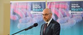 Mężczyzna w czarnym garniturze i w okularach stoi przy mównicy i mówi do mikrofonu na stojaku, za nim przed żółtą ścianą dwa rozwinięte niebiesko-różowe banery z napisami Konferencja „Elbląg PUBLIC HEALTH”.