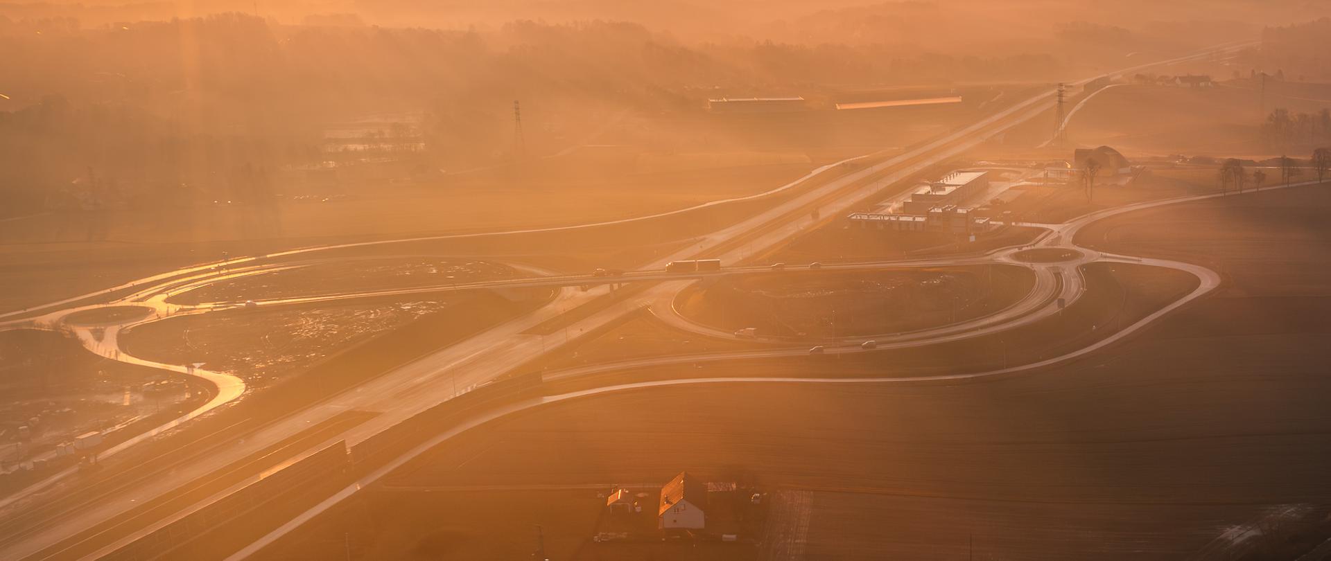 Węzeł drogowy Stara Wieś wraz z Obwodem Utrzymania Drogi z lotu ptaka w ciepłym, złocistym świetle wschodzącego słońca. Wokół węzła rozciągają się pola uprawne i nieliczne zabudowania, w tym pojedynczy dom w dolnej części kadru. Całość otacza lekko zamglony, pagórkowaty teren, nadający zdjęciu malowniczy i spokojny charakter.