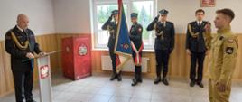 Uroczystość ślubowania strażaka w siedzibie Państwowej Straży Pożarnej. Po prawej stronie strażak w ubraniu służbowym składa ślubowanie z uniesioną prawą ręką, po lewej komendant odczytuje rotę ślubowania. W tle poczet sztandarowy oraz strażacy w umundurowaniu galowym