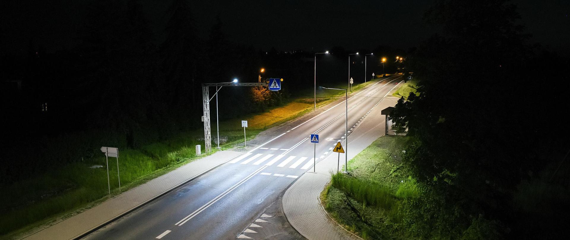 Widok z góry na przejście dla pieszych w Radymnie. Pora nocna, świeci oświetlenie drogowe. Z prawej strony widać zatokę autobusową i przystanek.