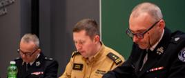 Zajęcie przedstawia pracującą komisję trzy osoby w mundurach strażackich podczas eliminacji powiatowych Ogólnopolskiego Turnieju Wiedzy Pożarniczej w powiecie gorlickim siedzących przy stole na sali Centrum Kształcenia zawodowego w Gorlicach i poprawiających testy 
