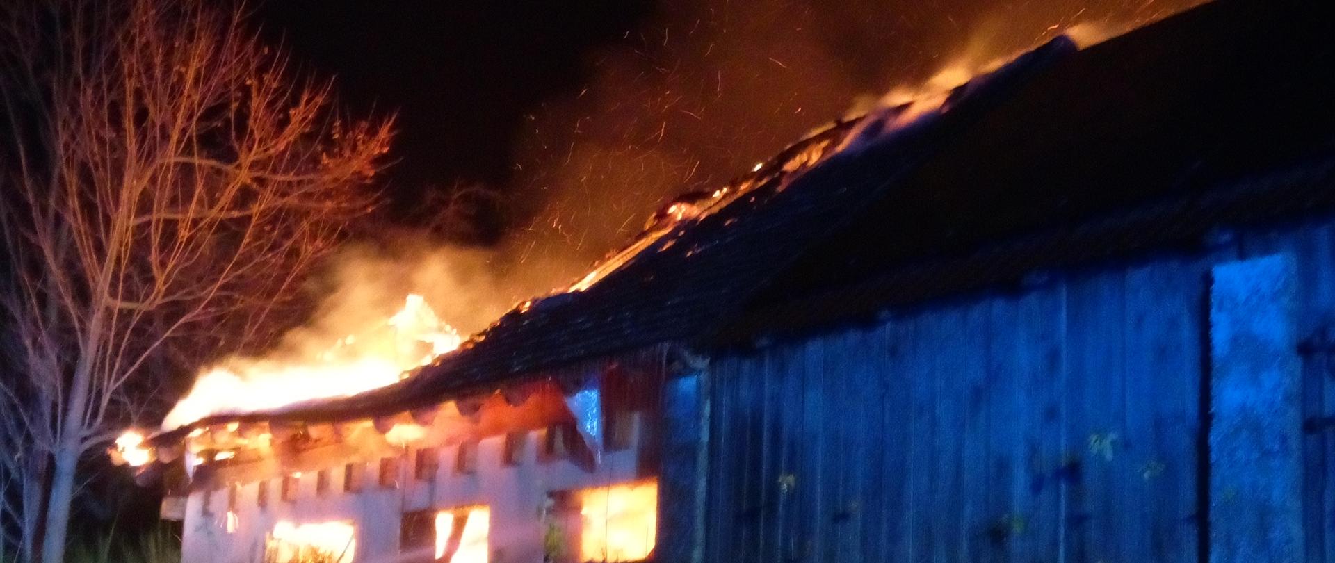 Zdjęcie przedstawia pożar budynku gospodarczego - dach budynku objęty jest ogniem. Z prawej strony widać strażaka podającego prąd gaśniczy.