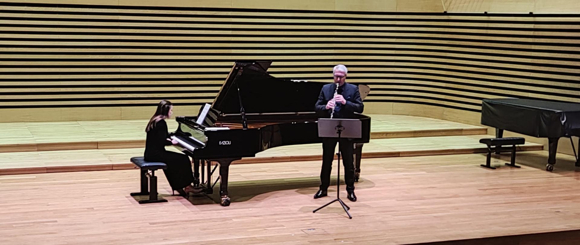 Mężczyzna stoi na estradzie sali koncertowej przy pulpicie i gra na klarnecie, za nim lekko po lewej stronie kobieta gra na fortepianie.