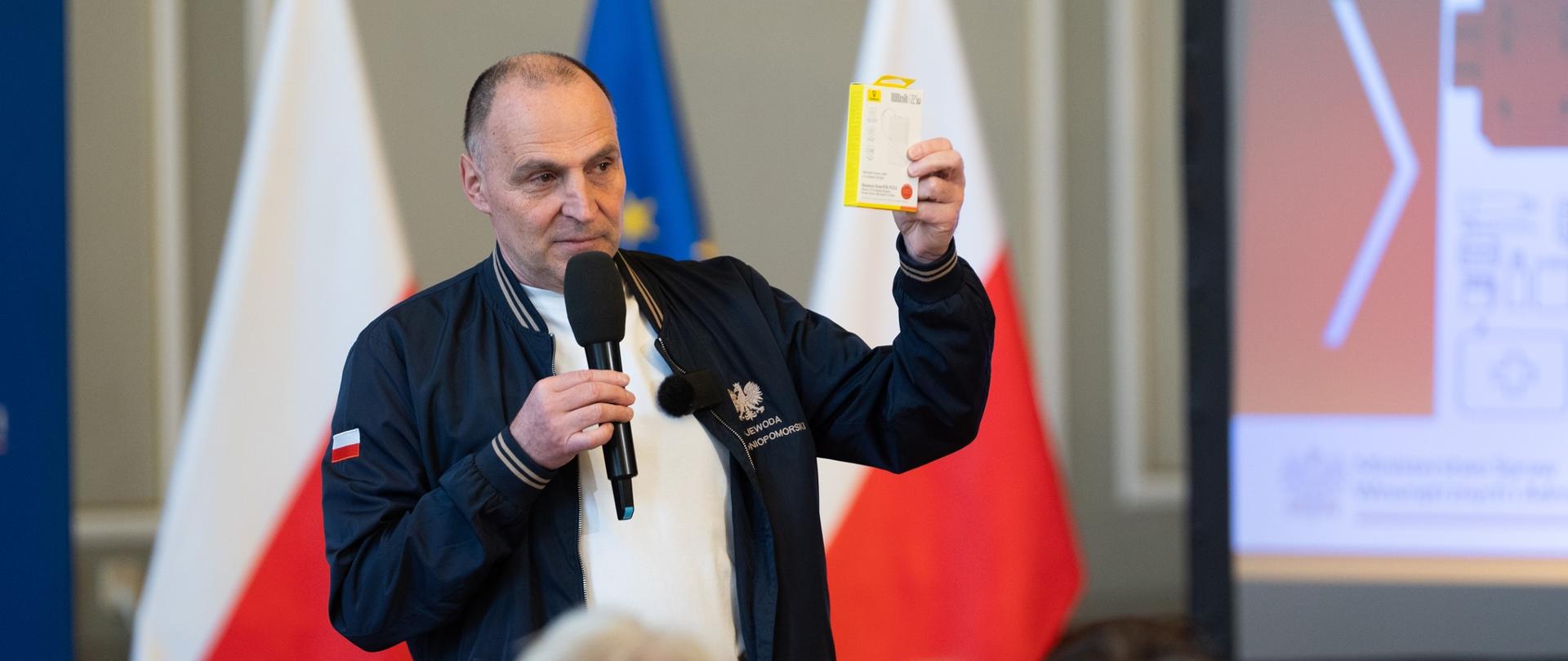 Zdjęcie przedstawia mężczyznę przemawiającego do publiczności podczas spotkania. Trzyma w jednej ręce mikrofon, a w drugiej unosi niewielkie pudełko, pokazując je uczestnikom –jako przykład lub element prezentacji. Ubrany jest w jasny strój z granatową kurtką.W tle widoczne są flagi Polski i Unii Europejskiej oraz fragment ekranu z wyświetlaną prezentacją. Na pierwszym planie, od tyłu, widać głowy słuchaczy siedzących na widowni. Całość sugeruje wystąpienie informacyjne lub szkoleniowe, podczas którego prelegent omawia konkretny przedmiot.