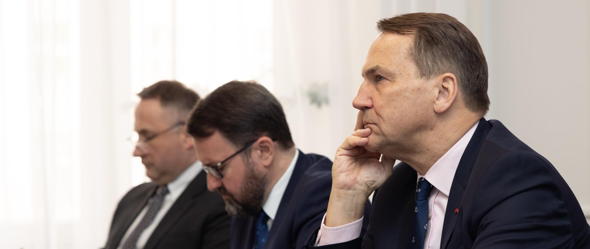 The photo shows three men. They are sitting at a table and taking notes. 