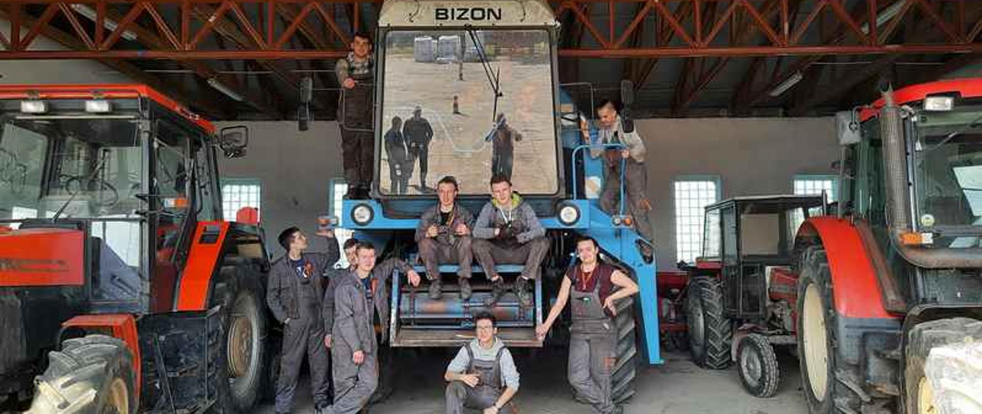 Garaż z maszynami rolniczymi. Naśrodkowym bizonie stoi i siedzi kilku chłopaków w strojach roboczych.