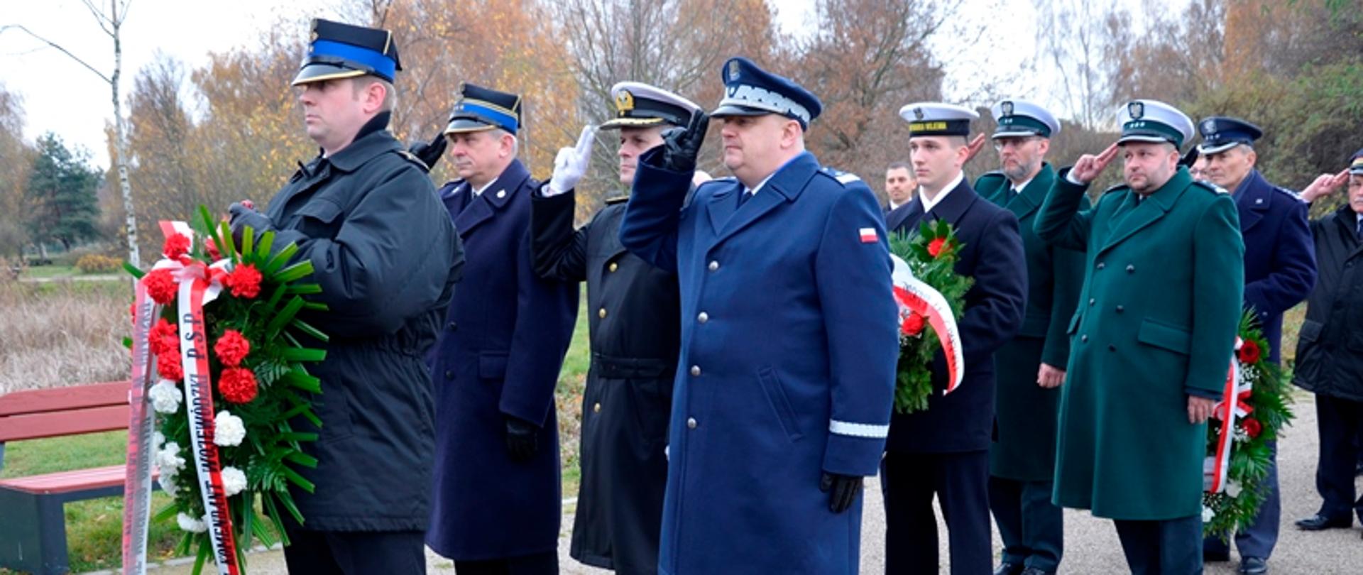 Przedstawiciele służb mundurowych składają wiązanki kwiaty, oddając honory.