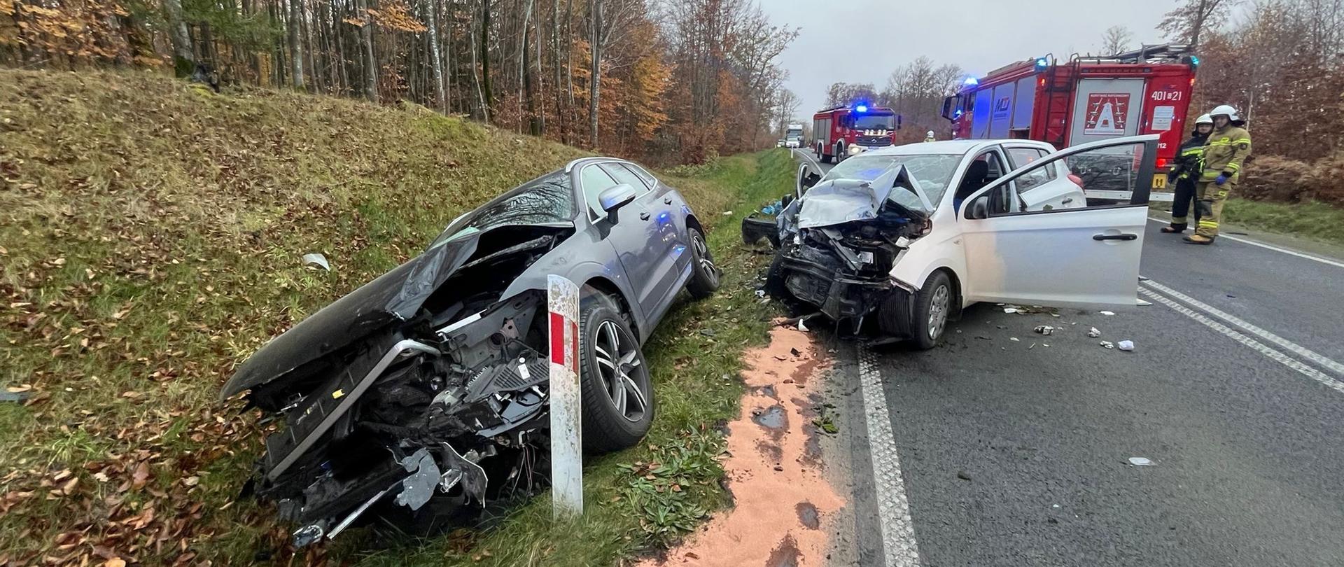 Dwa rozbite samochody. Jeden w rowie, a drugi na jezdni. W tle strażacy i pojazdy pożarnicze. 