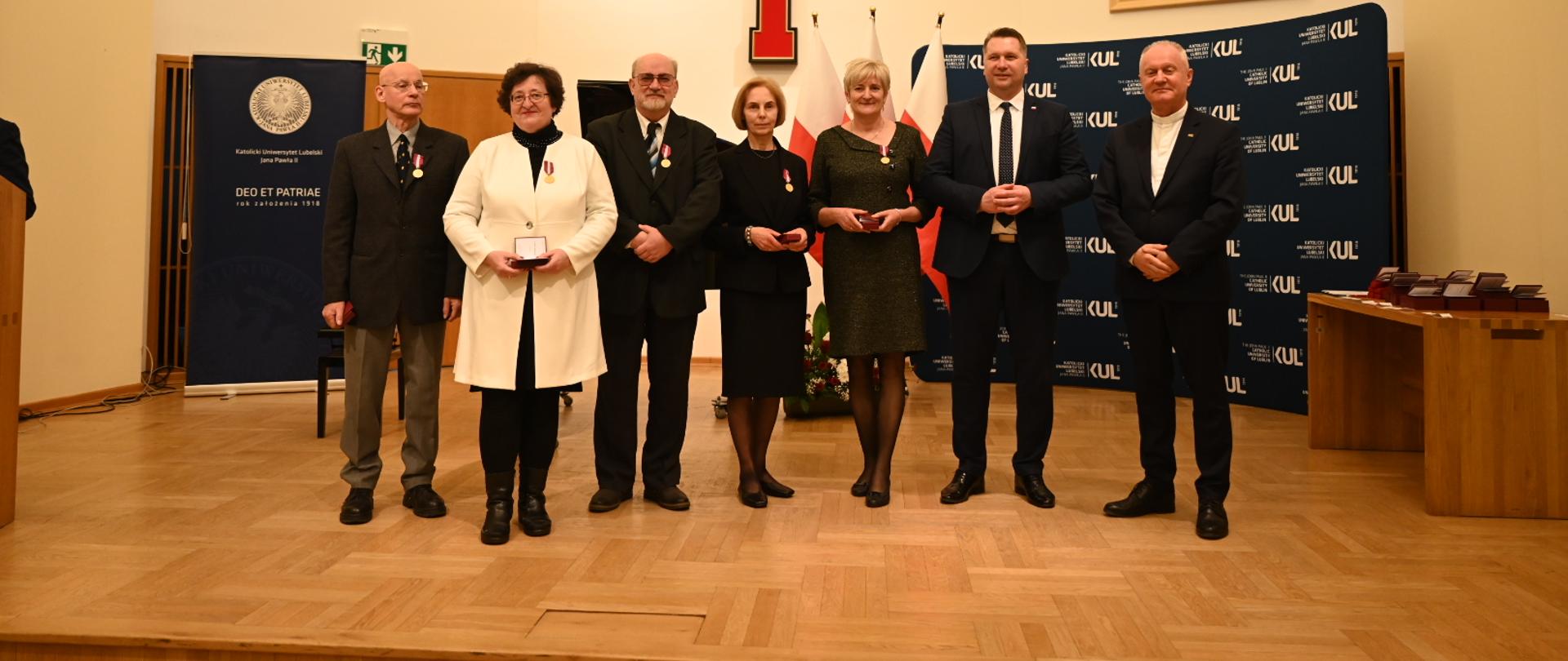 Grupa ludzi, czterech mężczyzn i trzy kobiety, stoi na drewnianej podłodze, za nimi na żółtej ścianie duży czerwony krzyż i ścianka z napisami KUL.
