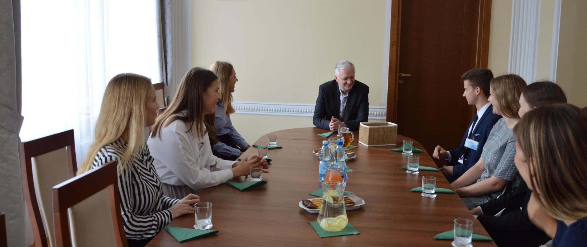 Minister siedzi za stołem, po obu jego stronach siedzą studenci