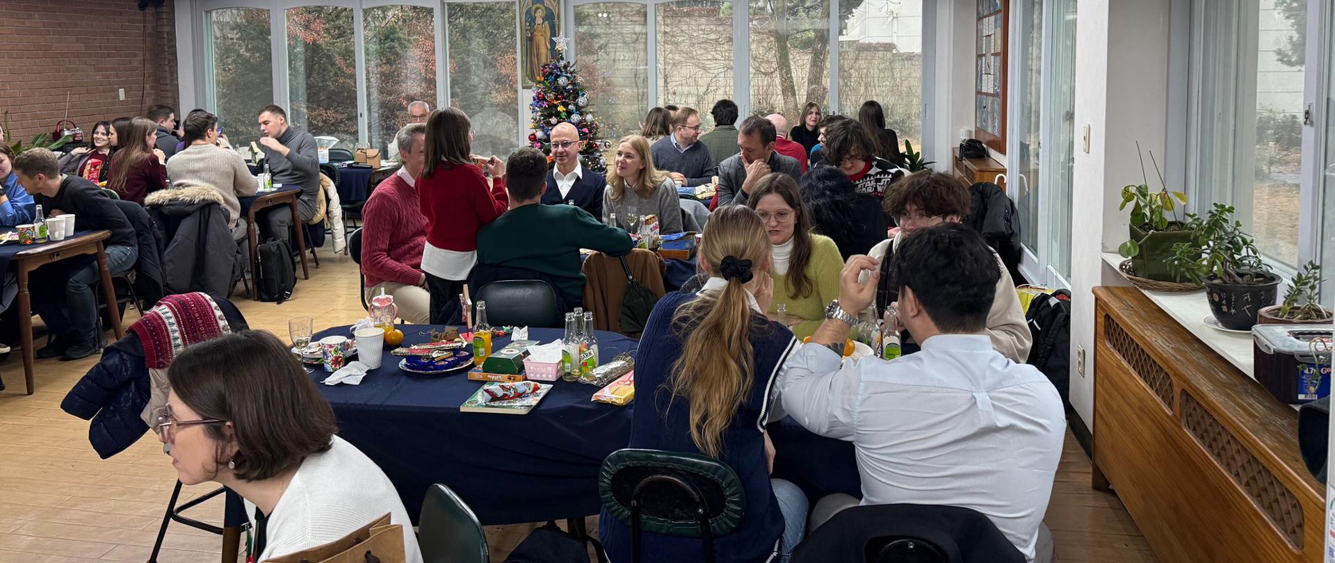 Zdjęcie przedstawia część uczestników spotkania bożonarodzeniowego Ambasady RP w Seulu dla polskich dzieci i Polaków mieszkających w Korei, które odbyło się 13 grudnia 2025 roku, siedzących przy stołach, na których rozłożone są polskie dania, słodycze i napoje.
