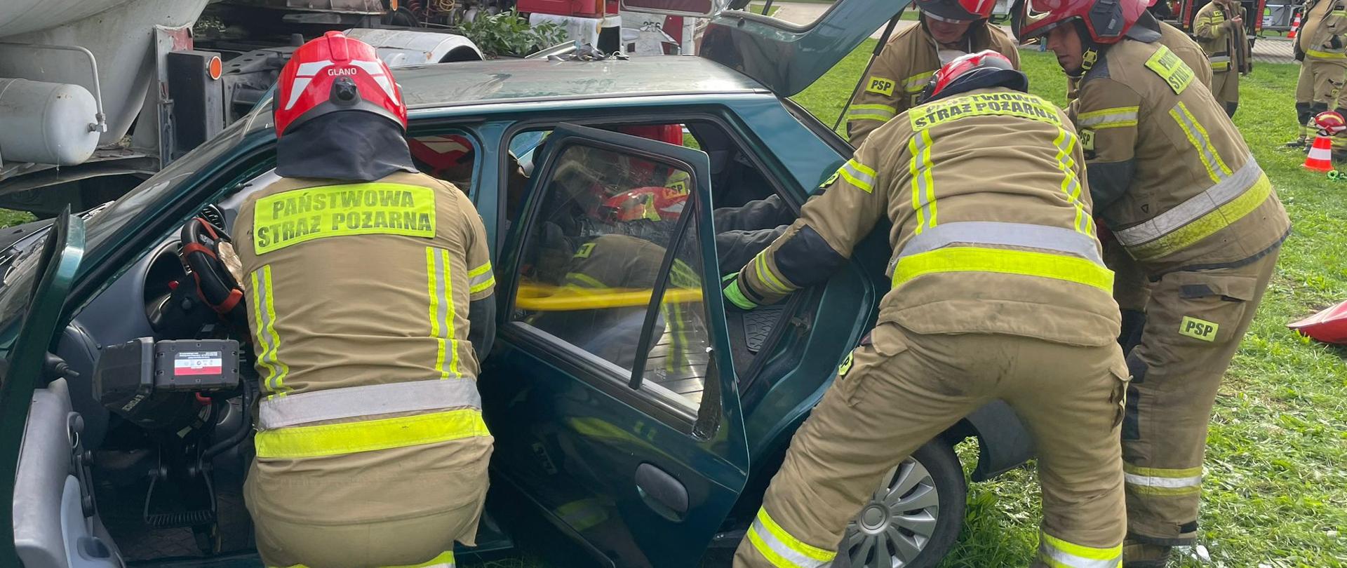 Strażacy PSP w umundurowaniu specjalnym i hełmach podczas szkolenia doskonalącego z ratownictwa technicznego w zakresie podstawowym realizowanego przez KSRG. Jeden z ratowników używa narzędzi hydraulicznych do cięcia karoserii uszkodzonego samochodu, a pozostali ewakuują pozoranta.