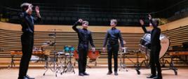 Czterech chłopców ubranych na czarno stoi na estradzie sali koncertowej PSM w Jastrzębiu-Zdroju i gestykuluje rękami. Za nimi stoją różne instrumenty perkusyjne.