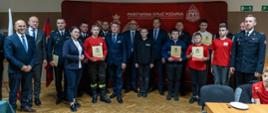 młodzieżowe drezyny pożarnicze MDP w towarzystwie parlamentarzystów władz wojewódzkich, powiatowych trzymają w ręku okolicznościowe promesy, wszyscy stoją na tle czerwonego baneru