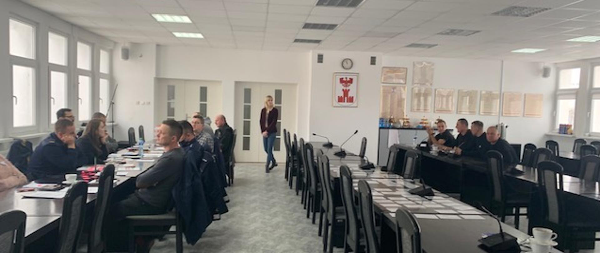 Zdjęcie przedstawia salę na której znajdują się czarne stoły przy których siedzi czterech mężczyzn po prawej stronie natomiast po lewej 8 mężczyzn i jena kobieta. Pomiędzy stołami stoi kobieta. Na ścianie widzimy zegar oraz herb Miasta Świdwin przedstawiający czerwonego orła będącego nad brama na białym tle.