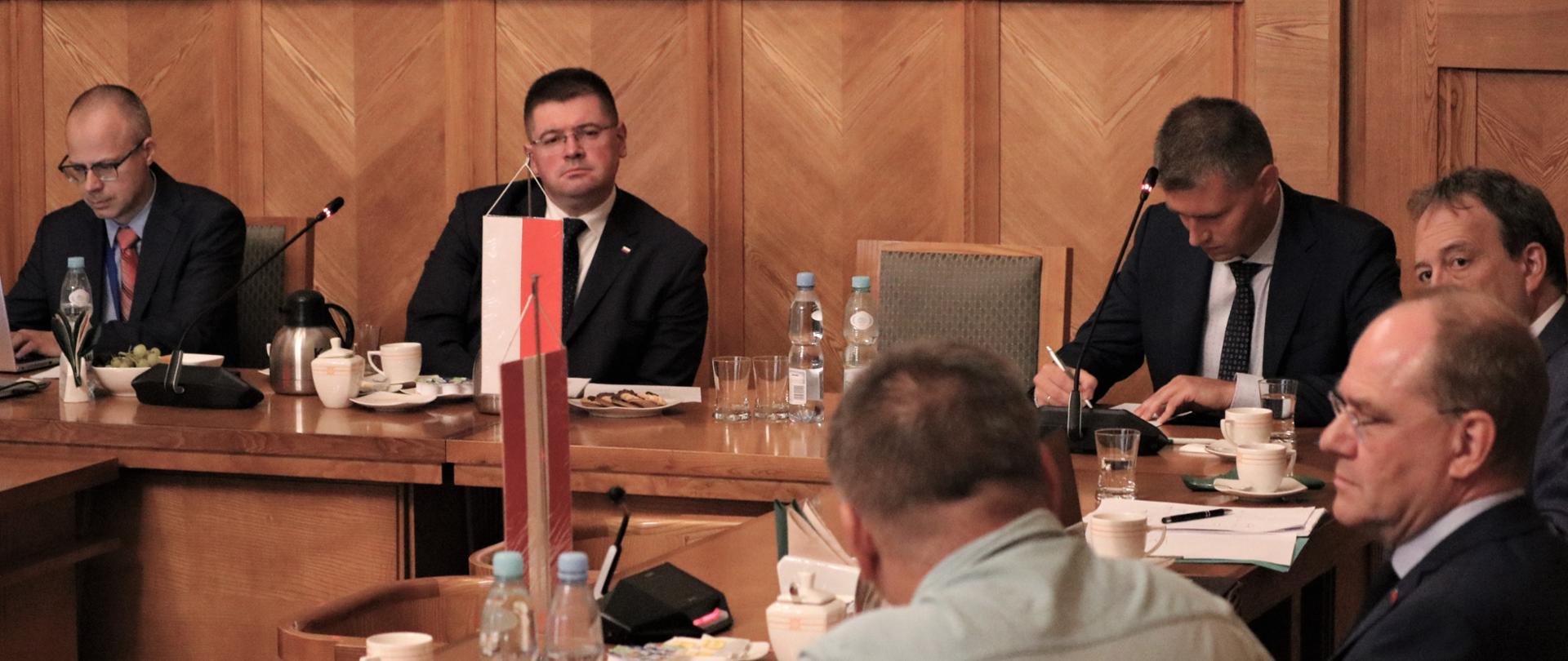 Sześciu mężczyzn siedzi przy stole, jeden pracuje na laptopie, drugi pisze na kartce, trzech słucha, jeden mówi. Na stole filiżanki, woda, ciastka i owoce