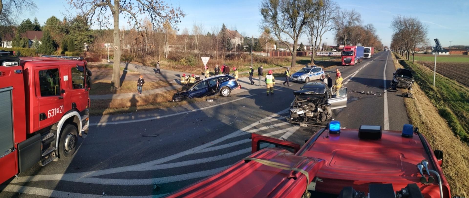 Zdjęcie wykonane z dachu samochodu strażackiego. Widok na drogę, na której stoją trzy rozbite samochody. Widać radiowóz policji, strażaków udzielających pomocy osobom poszkodowanym. W oddali stojące w korku samochody ciężarowe.