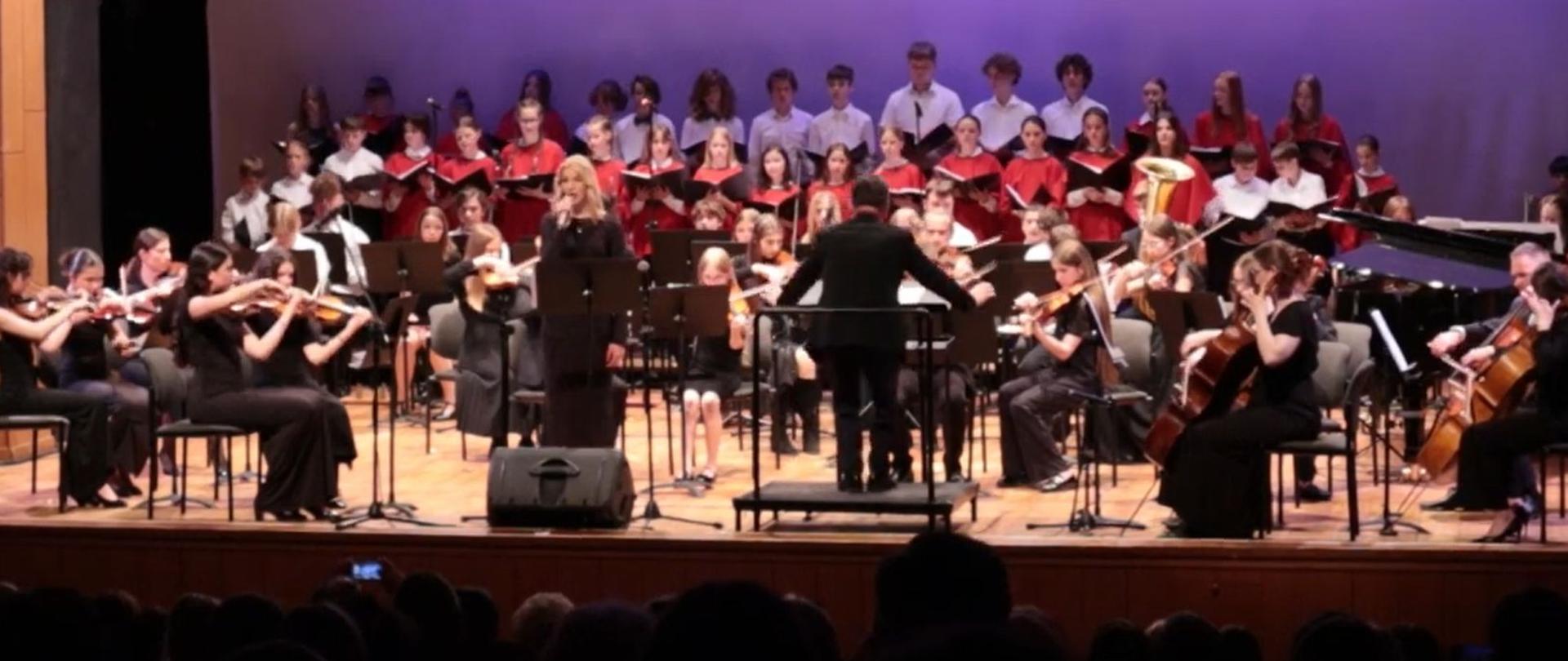 Zdjęcie kolorowe. Orkiestra i chór na scenie. Z przodu solistka z mikrofonem.
