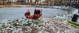 Na zdjęciu widoczna jest symulacja akcji ratowniczej na zamarzniętym zbiorniku wodnym. Strażacy w ubraniach do ratownictwa lodowego ewakuują poszkodowanego przy użyciu sanek lodowych i lin asekuracyjnych. Działania prowadzone są z zachowaniem zasad bezpieczeństwa i odpowiedniego zabezpieczenia ratowników. W tle zgromadzeni mieszkańcy obserwują przebieg ćwiczeń z brzegu zbiornika. Zdjęcie dokumentuje praktyczny pokaz ratownictwa lodowego.