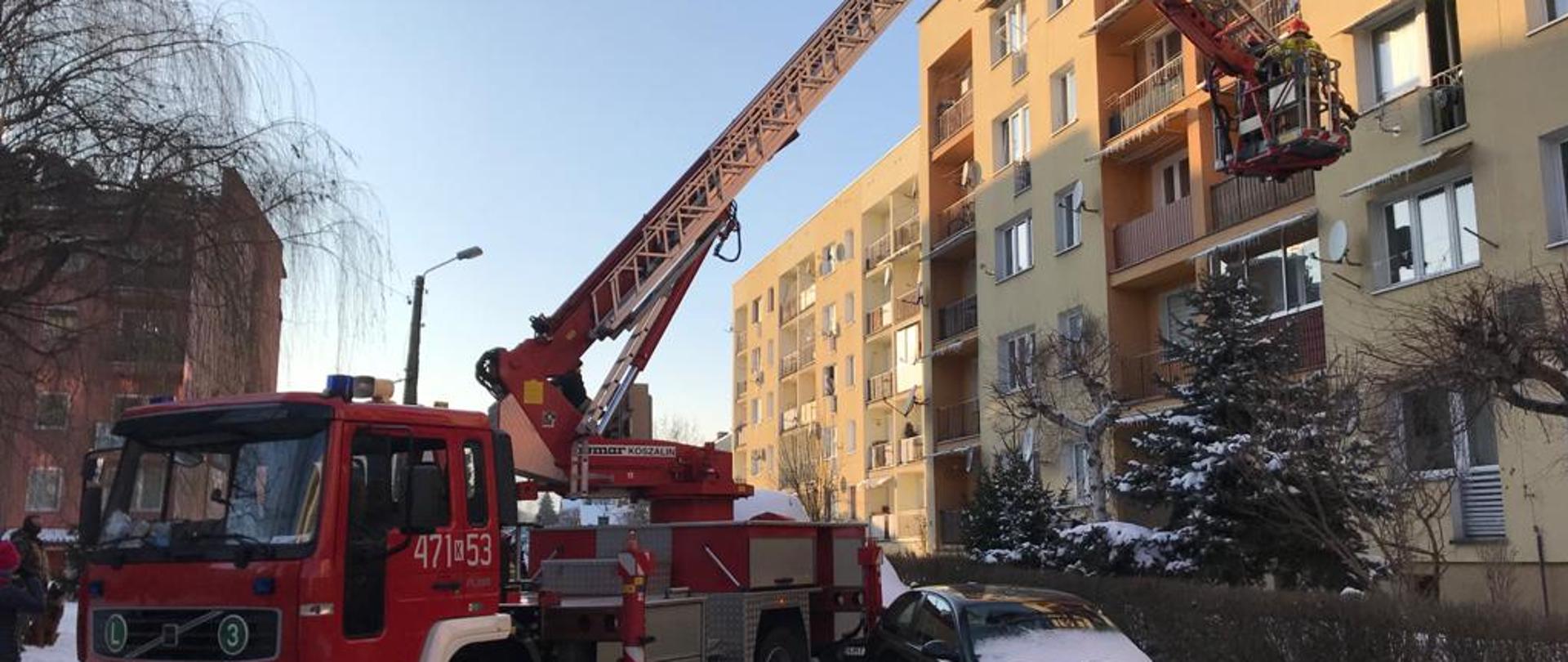 Na zdjęciu widoczny jest rozłożony podnośnik hydrauliczny. Ramię wysięgnika zbliża się do drzwi balkonowych. W koszu znajdują się ratownicy. Obok chodnika widoczny zaparkowany samochód osobowy w kolorze czarnym.