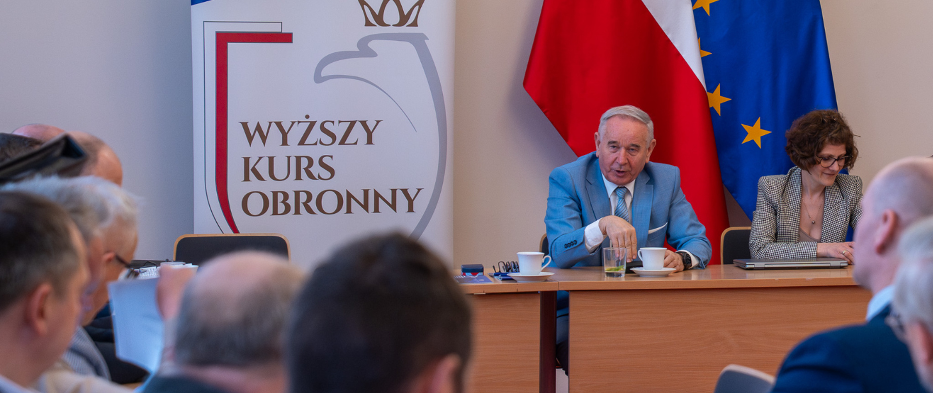 Spotkanie szkoleniowe z udziałem Wojewody Kujawsko-Pomorskiego, Michała Sztybla. W tle flagi Polski i Unii Europejskiej oraz roll-upy organizatorów. W tle widoczna jest prezentacja na ścianie. 