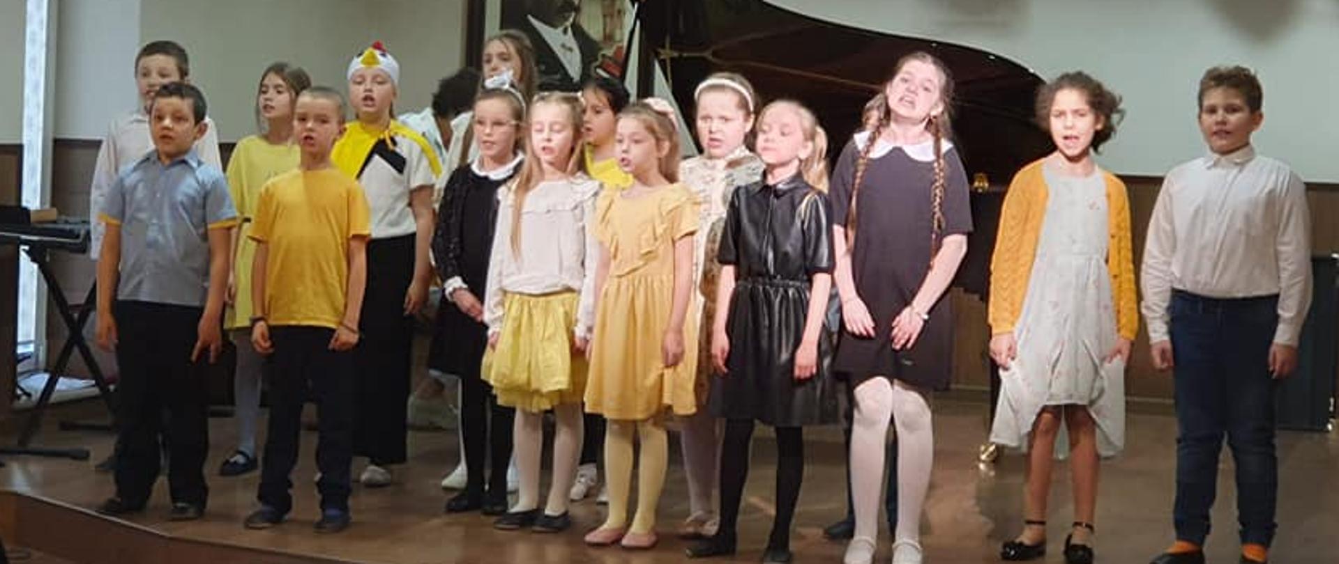 Na środku sceny stoją śpiewający w chórku uczniowie klasy pierwszej, Podłoga sceny w kolorze brązy, z lewej strony fragment okna, przy którym stoi statyw na nuty i instrument dzwonki, za uczniami widać otwartą klapę fortepianu. Z lewej strony mikrofon. Przed chórkiem stopień na scenę i fragmenty oparć krzeseł. Na suficie okrągłe świecące lampy.