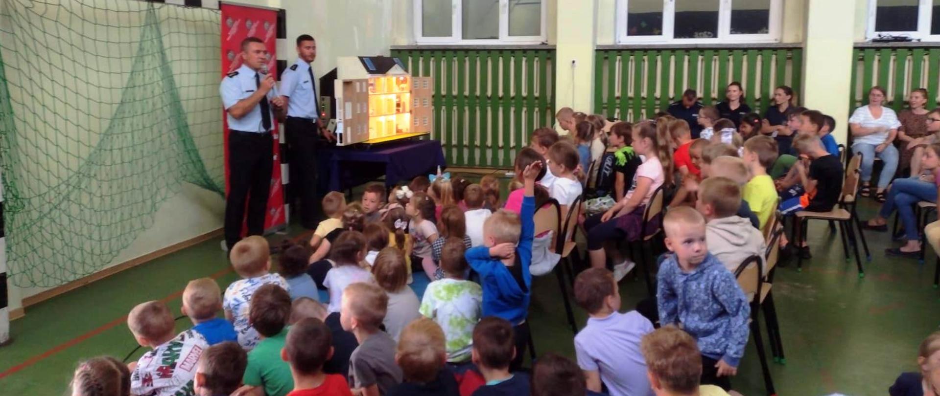 Zdjęcie przedstawia strażaków którzy przy wykorzystaniu mobilnego symulatora zagrożeń pożarowych omawiają dzieciom na sali gimnastycznej zagrożenia występujące w domu. Dzieci siedzą i obserwują strażaków. 