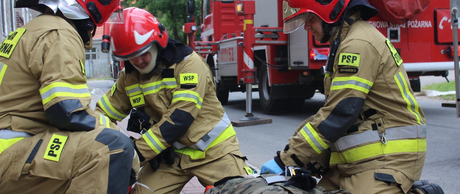 Na zdjęciu trzech strażaków w mundurach bojowych i hełmach opatrują leżącego na chodniku żołnierza. W tle samochód strażacki.