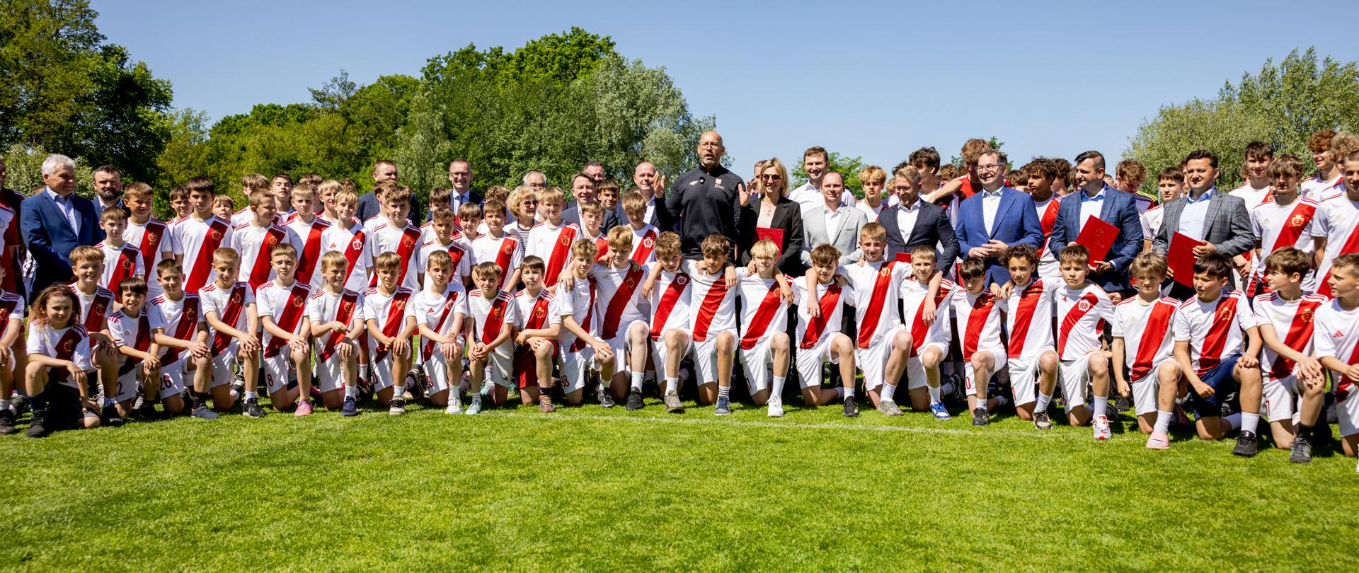 Zbiorowe zdjęcie młodych zawodników Gdańskiego Klubu Sportowego Gedania 1922 i ministra sportu i turystyki Jakuba Rutnickiego. Zdjęcie zrobione na boisku piłkarskim. 