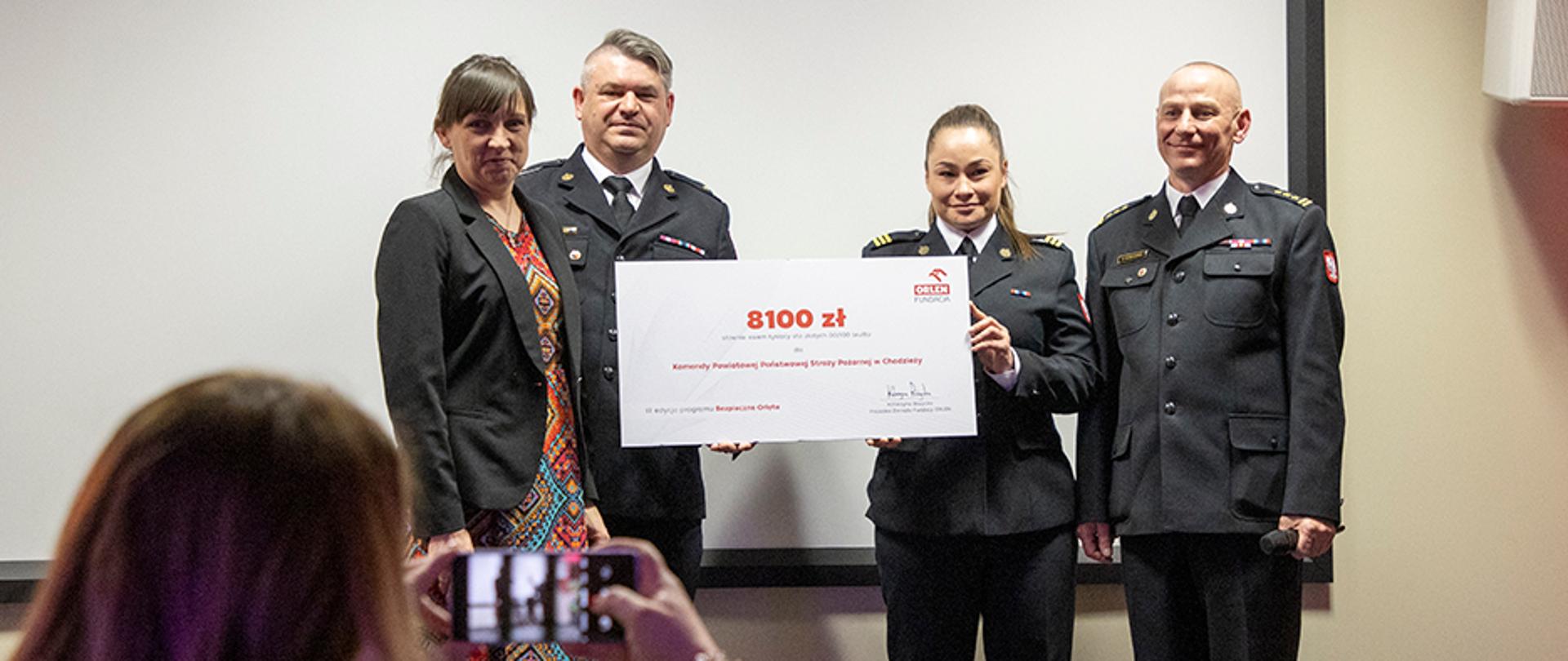 Kobieta oraz dwóch mężczyzn w strażackich mundurach oraz kobieta we wzorzystej sukience i czarnej marynarce prezentujący czek z wyróżnioną kwotą osiemtysięcysto złotych. Na pierwszym planie w lewym dolnym rogu kobieta robiąca zdjęcie smatrfonem