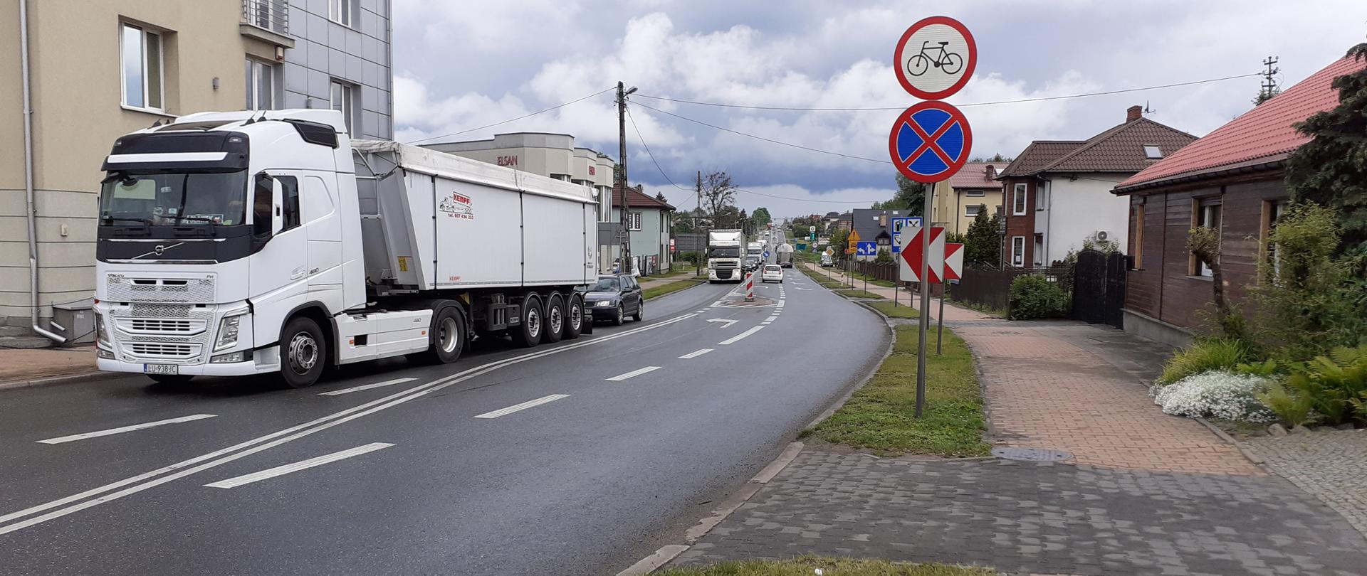 Na zdjęciu widać fragment ulicy w miejscowości o zabudowie mieszkalnej.
Na jezdni znajduje się duży biały ciągnik siodłowy z naczepą (ciężarówka), jadący w kierunku widza. Za nim widoczny jest kolejny pojazd ciężarowy w oddali.
Droga asfaltową ulicą z dwoma pasami ruchu (po jednym w każdą stronę), z wyraźnymi liniami rozdzielającymi pasy. Nawierzchnia wygląda na mokrą, co sugeruje niedawny deszcz. Po prawej stronie stoją dwa znaki:
Zakaz wjazdu rowerów (czerwone koło z symbolem roweru).
Zakaz zatrzymywania się (niebieskie koło z czerwoną obwódką i przekreśleniem).
Po lewej stronie znajduje się budynek wielopiętrowy w jasnym kolorze, a po prawej domy jednorodzinne z czerwonymi dachami i ogródkami. Widać chodnik z kostki brukowej oraz fragment trawnika.
Droga prowadzi w głąb miejscowości, widać kolejne budynki i pojazdy. Na niebie są ciemne chmury, co sugeruje pochmurną pogodę lub nadchodzącą burzę.