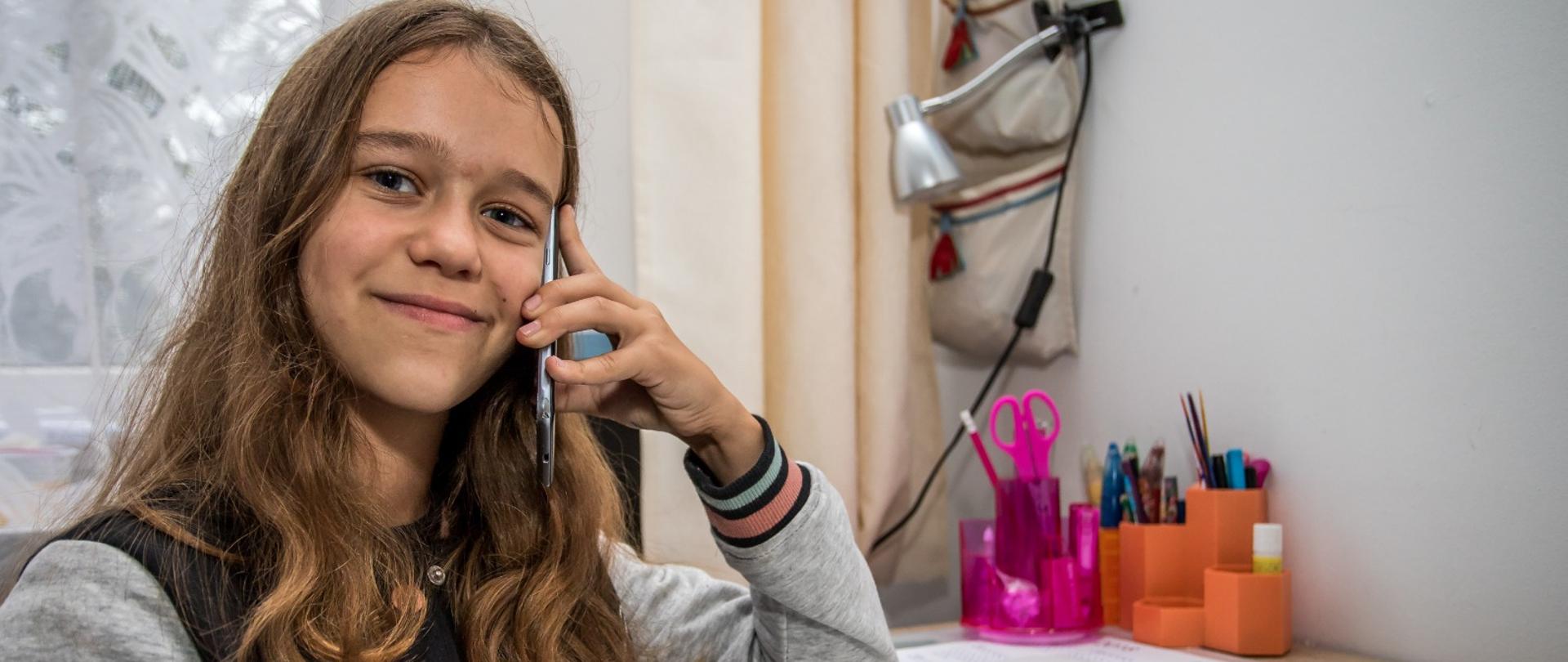 Na zdjęciu uśmiechnięta dziewczynka w wieku szkolnym, trzymająca w prawej dłoni słuchawkę telefonu komórkowego. Dziewczynka siedzi przy biurku, na którym znajduje się różowy i pomarańczowy przybornik z długopisami i kredkami. W tle widać okno zasłonięte firanką oraz lampkę. 