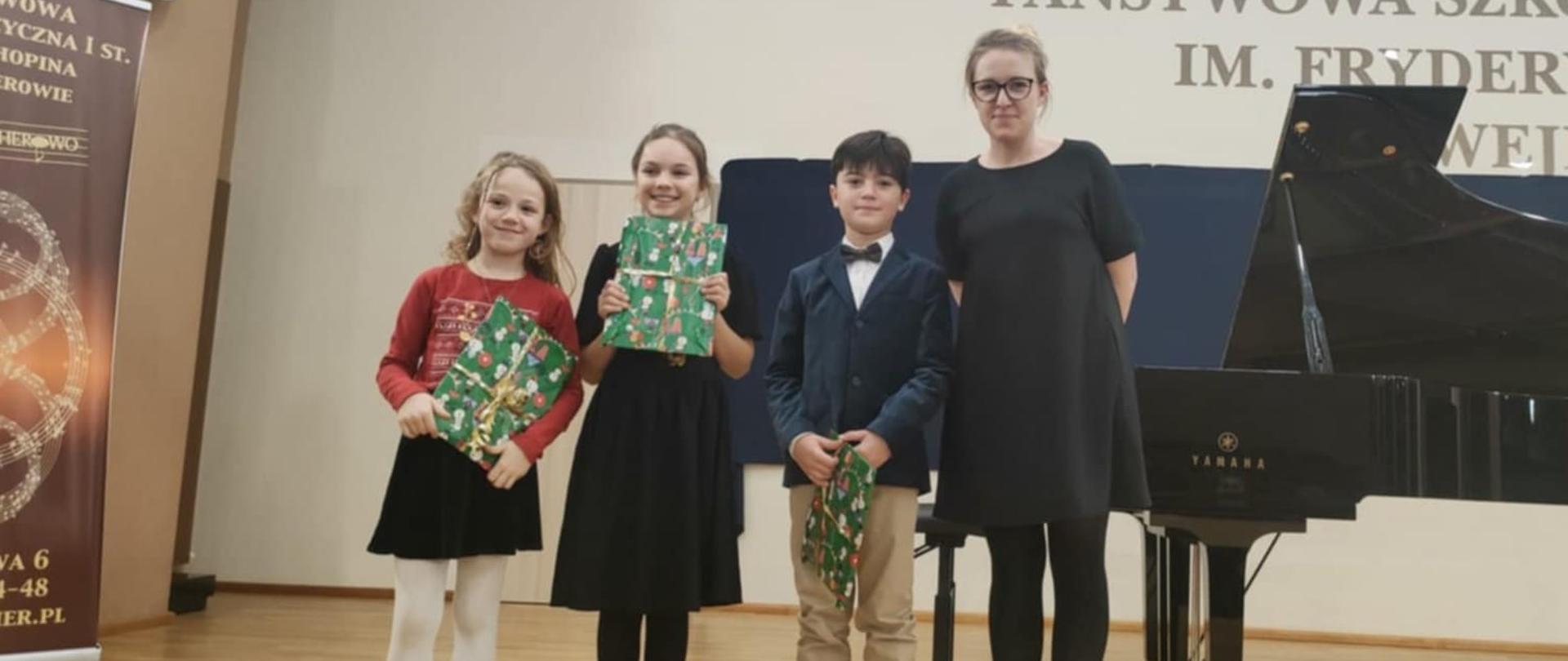 Na scenie sali koncertowej pozuje do zdjęcia pani Anna Zazula-Chajęcka wraz z trójką swoich uczniów.