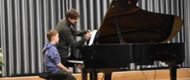 na scenie auli PSM, podczas warsztatów pianistycznych, widnieje uczeń i nauczyciel, którzy pracują nad utworem muzycznym. Uczeń siedzi przy fortepianie, nauczyciel wprowadza uwagi do tekstu muzycznego. Kolorystyka zdjęcia jest brązowo-szaro-niebiesko- biało-czarna. 