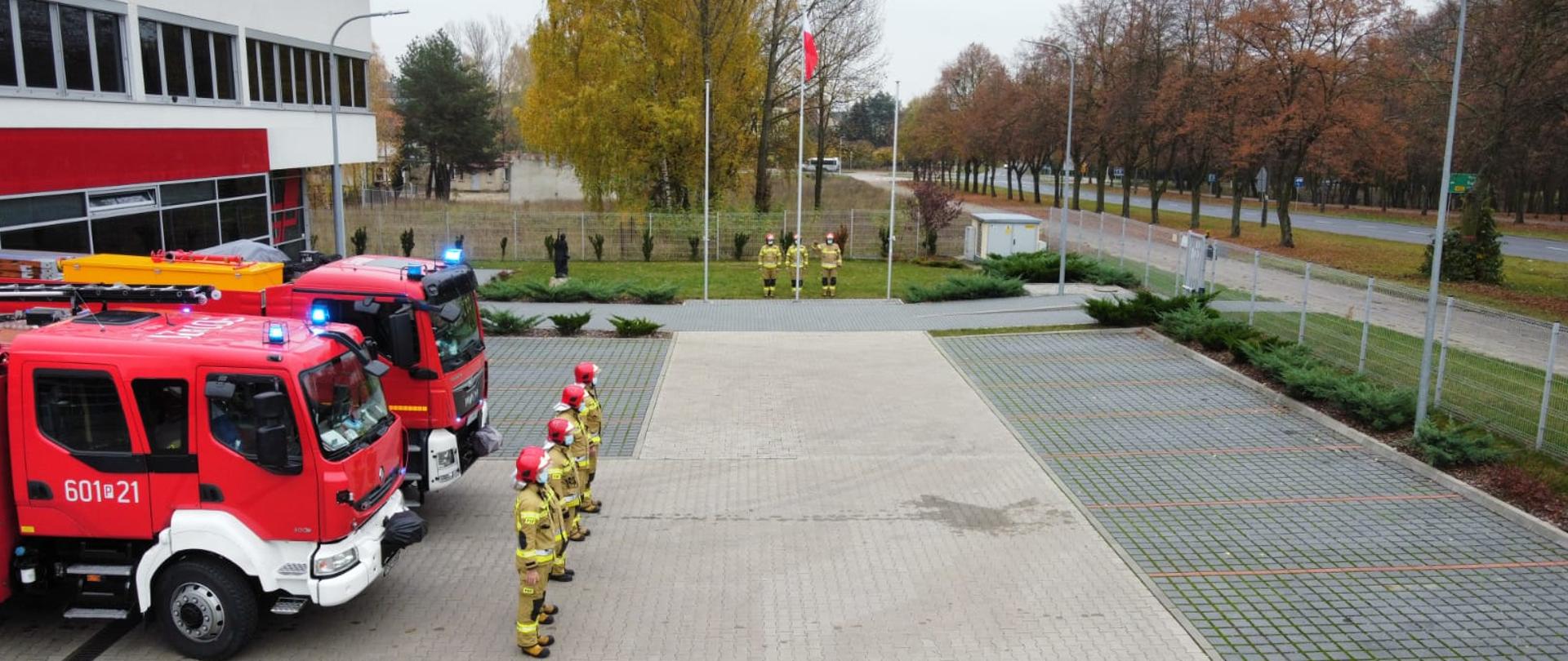 Zdjęcie pokazuje samochody ratowniczo gaśnicze zmianę służbową oraz poczet flagowy przy maszcie na który zostaje podniesiona flaga państwowa
