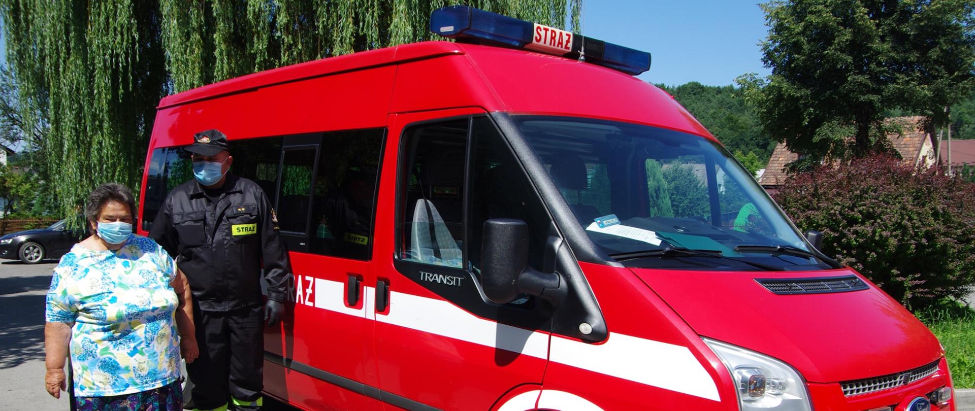 Widok z przodu. Strażak PSP w czarnym ubraniu koszarowym i maseczce ochronnej stoi przy samochodzie strażackim stoi obok starszej kobiety w maseczce ochronnej. 