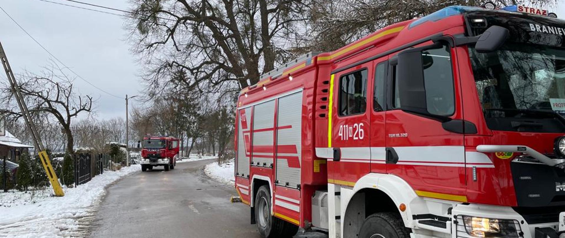Na drodze stoją dwa samochody czerwone strażackie. Pobocza białe od śniegu. W oddali drzewa.