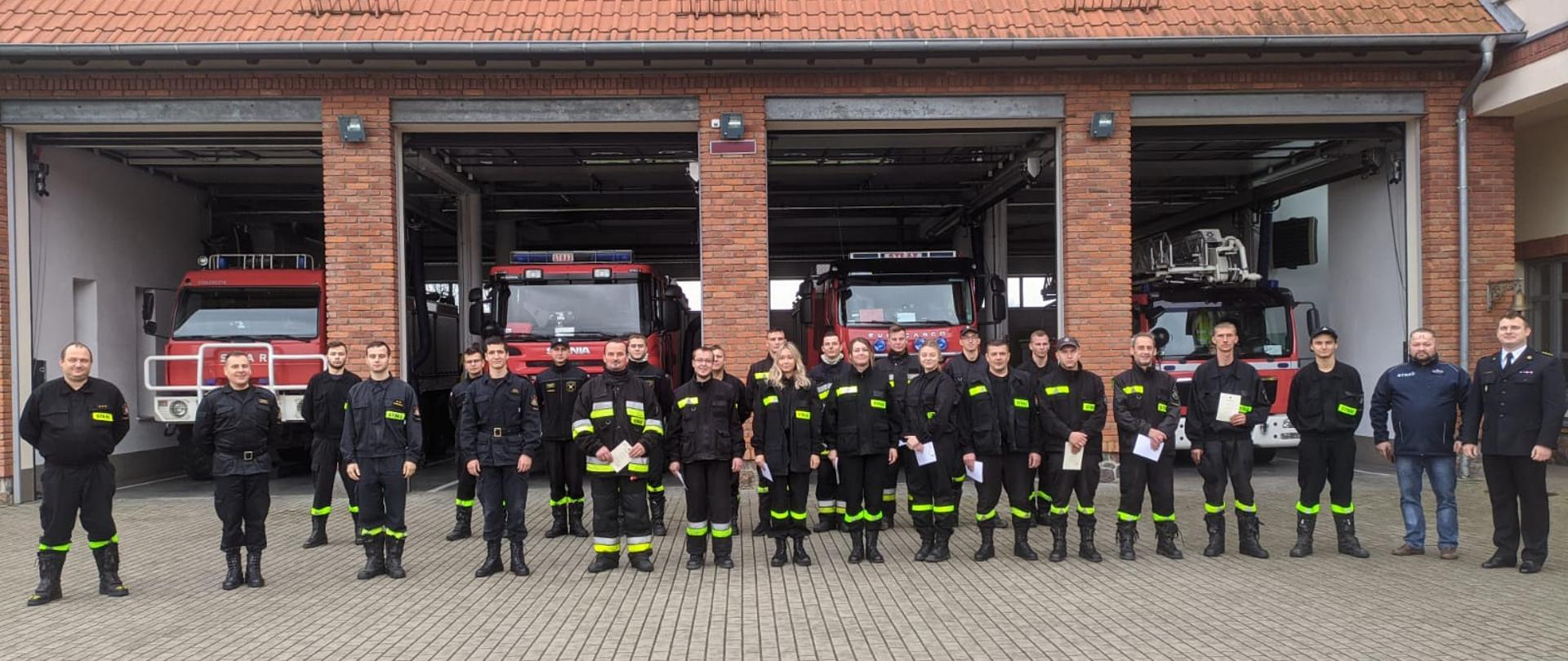 Zdjęcie przedstawia m.in. absolwentów szkolenia podstawowego strażaków ratowników OSP z zaświadczeniami o jego ukończeniu oraz zespół egzaminacyjny, na tle pojazdów pożarniczych stacjonujących w garażu Komendy Powiatowej Państwowej Straży Pożarnej w Gołdapi