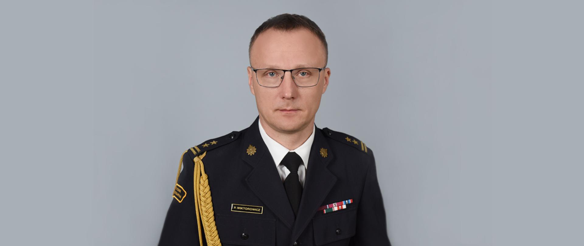 Zdjęcie to oficjalny portret mężczyzny w mundurze wyjściowym Państwowej Straży Pożarnej, przedstawiony na jednolitym, szarym tle.
Postać
• Mężczyzna ma krótkie, ciemne włosy, nosi okulary w ciemnych oprawkach i patrzy bezpośrednio w stronę obiektywu z poważnym wyrazem twarzy.
• Jest ubrany w ciemnogranatowy mundur wyjściowy, białą koszulę oraz czarny krawat.
Detale munduru
Mundur jest bogato zdobiony insygniami świadczącymi o stopniu i zasługach:
• Akselbanty: Na prawym ramieniu widoczny jest gruby, pleciony sznur galowy (oficerski) w kolorze złotym.
• Pagony: Na ramionach znajdują się pagony z dwiema złotymi gwiazdkami i dwiema belkami, co odpowiada stopniowi brygadiera.
• Baretki: Nad lewą kieszenią przypięte są baretki odznaczeń państwowych i resortowych w różnych kolorach.
• Tabliczka imienna: Nad prawą kieszenią znajduje się czarna plakietka ze złotym napisem: P. WIKTOROWICZ.
• Emblematy: Na kołnierzu widoczne są złote emblematy korpusu straży pożarnej (ogniki). Na prawym rękawie znajduje się częściowo widoczna naszywka z napisem „PAŃSTWOWA STRAŻ POŻARNA”.
Zdjęcie charakteryzuje się profesjonalnym oświetleniem, jest ostre i ma wyważoną kompozycję typu popiersie.
